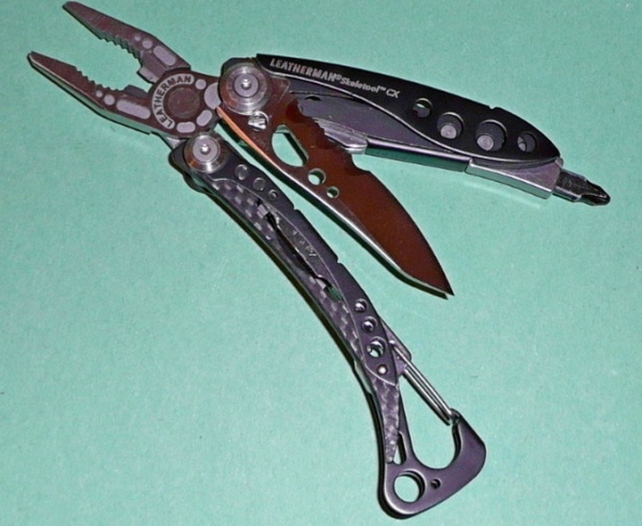 A Leatherman Skeletool CX multi-tool with its pliers, knife and bit driver opened, lying on a green surface